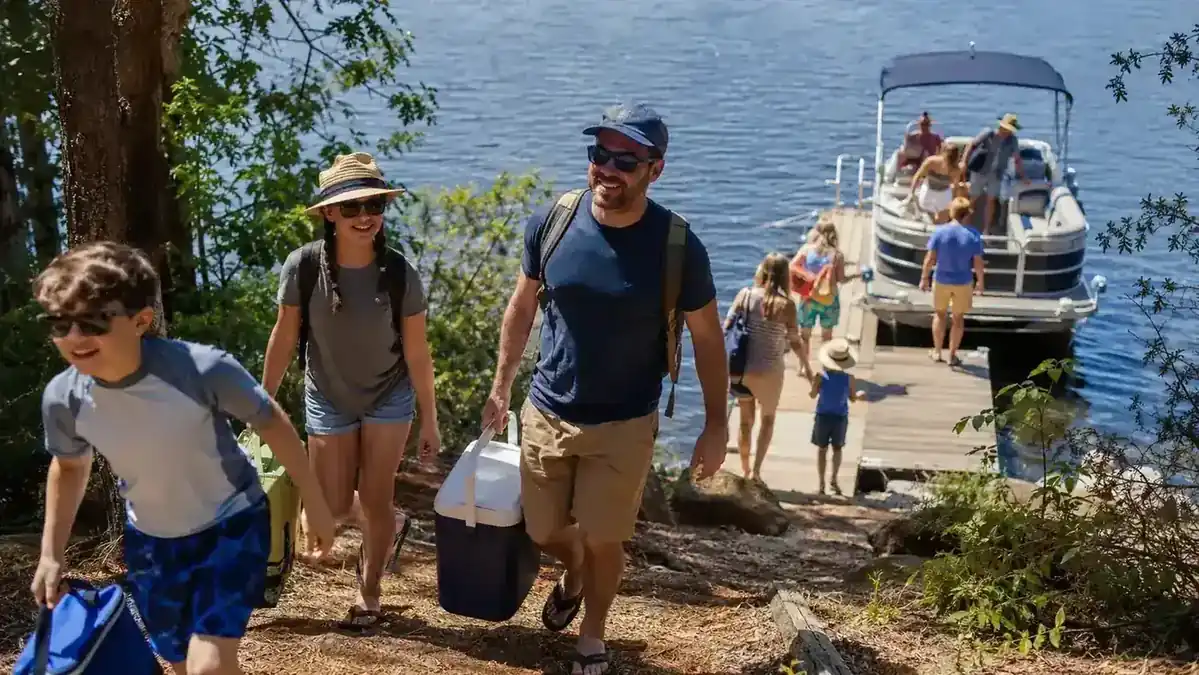 Family hauling coolers and gear down toward a dock and pontoon boat at a lake house