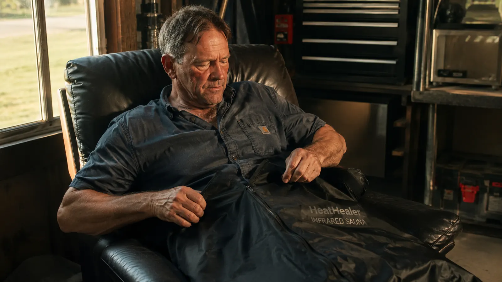 Man in his fifties in work clothes sitting in a recliner unzipping a black infrared sauna blanket in a garage in the evening
