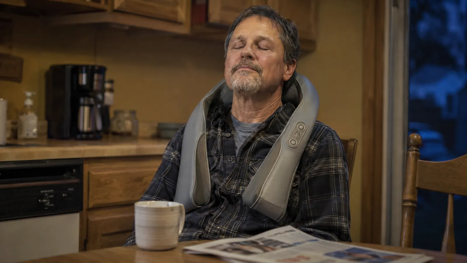 Man sitting at a kitchen table in the evening with a shiatsu neck massager draped over his shoulders, eyes closed